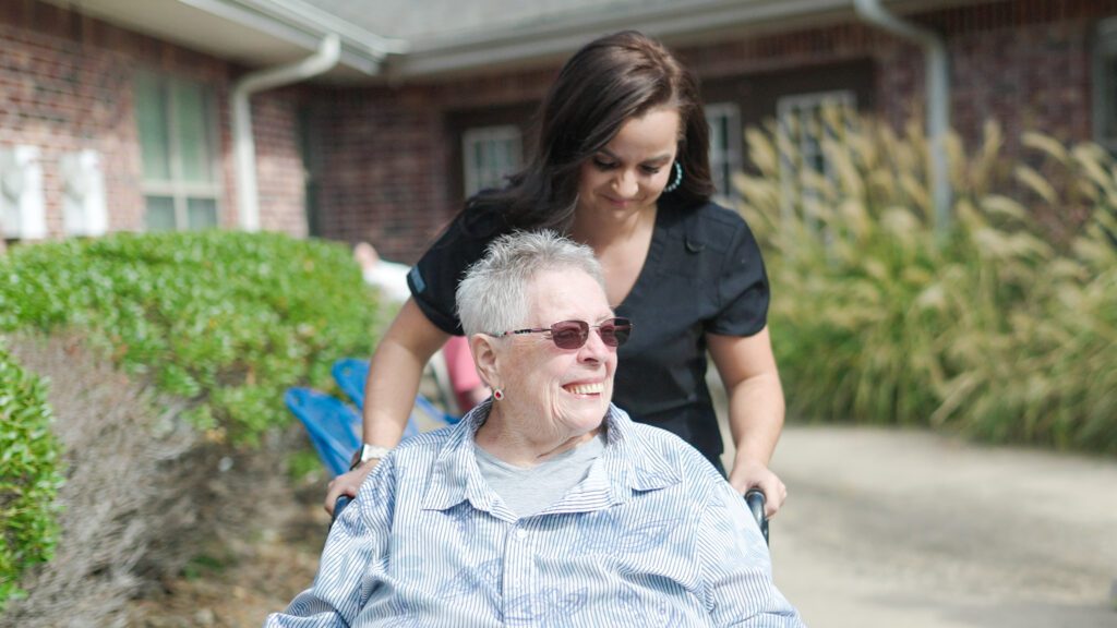 A smiling woman in a wheelchair is being pushed by a nurse at a skilled nursing facility