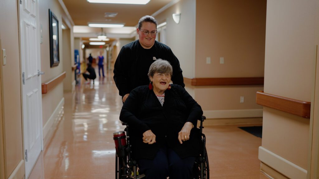 A woman in a wheel chair is smiling and being pushed by a smiling nurse at a long term care facility